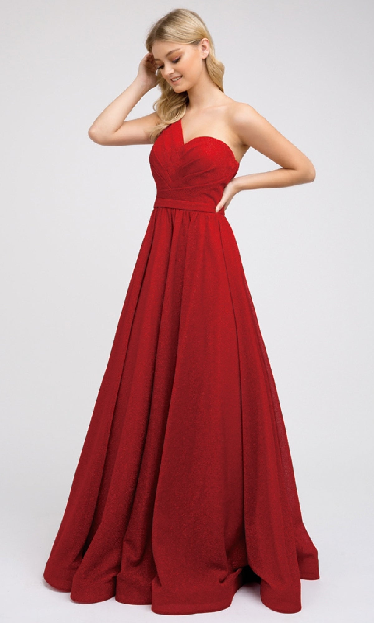 A woman in the Juliet 205 Long Prom Dress—an elegant, floor-length, one-shoulder red gown—smiles and touches her hair against a plain light background.
